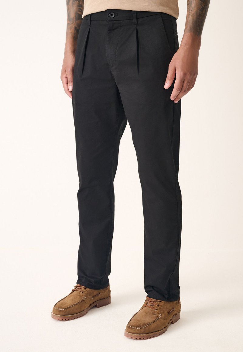 Black trousers with pleats, made of smooth fabric, feature a classic cut and a single button closure at the waist. Worn with brown suede shoes.