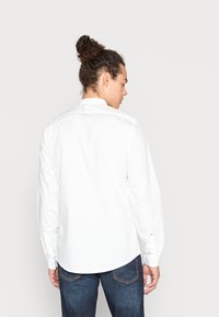 Camicia bianca a maniche lunghe in cotone, con colletto a pistagna e orlo curvo, abbinata a jeans in denim blu scuro.