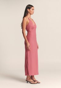 Ilga, prigludusi suknelė iš minkštos rožinės medžiagos, turinti gilų V formos iškirptę, plonas petnešėles ir subtilias šonines detales, derinta su juodais dirželiais puoštomis avalinės.