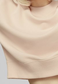 Felpa corta di un rosa chiaro con scollo rotondo e orlo a costine. Il tessuto appare morbido con una texture liscia e dettagli minimi.