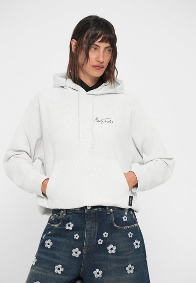 Siv hoodie z sprednjim žepom in vrvico, z črnim logotipom. V kombinaciji z temno modrimi kavbojkami, okrašenimi s cvetličnim vezenjem.