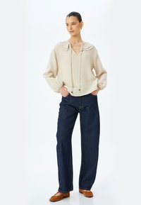 Beige, lichtgewicht blouse met lange mouwen, een grote kraag, een koordsluiting bij de hals en pofmouwen, gecombineerd met donkere, wijde denim jeans en bruine schoenen.
