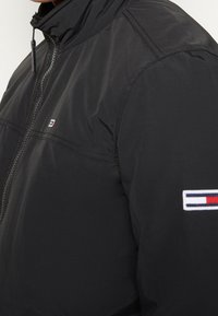 Giacca nera con zip caratterizzata da un colletto alto, tessuto texturizzato e un patch con logo sulla manica, con accenti in rosso, bianco e blu.
