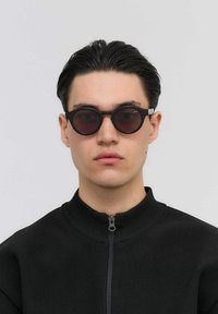 Musta pyöreät aurinkolasit tummilla linsseillä, metallisilla yksityiskohdilla ja paksulla kehyställä. Mallilla on päällään musta vetoketjullinen takki jossa on uritettu pinta.