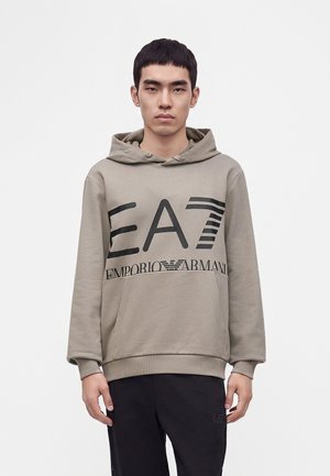 Jonge man die een beige hoodie draagt met een groot zwart EA7 Emporio Armani-logo en een zwarte broek, staande tegen een effen witte achtergrond.