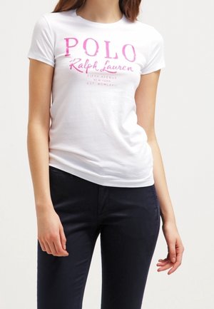 Vrouw draagt een wit aansluitend T-shirt met roze "Polo Ralph Lauren" tekst en donkerblauwe broek, staand tegen een effen achtergrond.