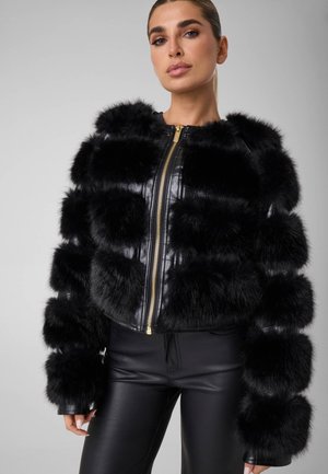 Modèle portant une veste en cuir noire bordée de fourrure, avec une fermeture éclair dorée, et un pantalon en cuir noir, sur un fond de studio gris.