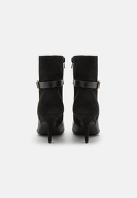 Bottines en daim noir avec un talon noir brillant, dotées d'une bride assortie avec des accessoires dorés. Texture lisse et silhouette élégante.