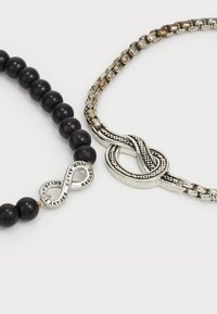 Due braccialetti: uno presenta perline tonde nere con un charm a forma di infinito argentato; l'altro ha una catena argentata texturizzata con un accento a forma di nodo.
