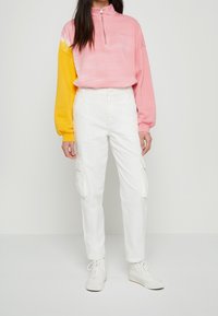 Rosa och gul zip-up tröja med puffiga ärmar, kombinerad med vita cargobyxor. Vita sneakers fullbordar outfiten.