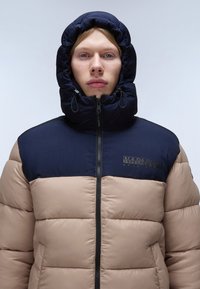 Giacca a vento con un design bicolore: parte superiore blu navy e parte inferiore beige. Cappuccio con cordini regolabili, chiusura lampo frontale e dettagli con marchio.