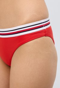 Κόκκινο μαγιό κάτω μέρος από λείο ύφασμα. Διαθέτει φαρδιά λευκή ελαστική μέση με κόκκινες και ναυτικές ρίγες. Κοντινή άποψη της πλευράς.