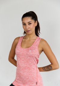 Vrouw met donker haar in een paardenstaart, draagt een roze sporttanktop en heeft een bloemtattoo op haar linkeronderarm, staande tegen een effen achtergrond.