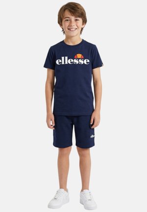 Garçon souriant portant un t-shirt ellesse bleu marine et un short assorti avec des baskets blanches, debout devant un fond clair uni.