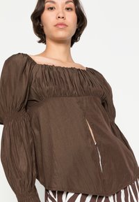 Top castanho com mangas bufantes, com decote franzido e fenda lateral, feito de tecido leve. A textura é suave com um brilho mínimo.