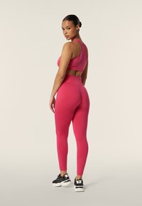Reggiseno sportivo rosa aderente e leggings, caratterizzati da una silhouette attillata e una texture liscia, abbinati a scarpe da ginnastica nere.