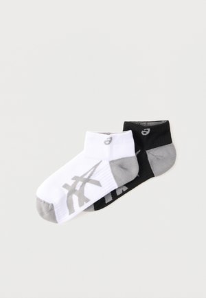 Jedna pár členkových ponožiek, jedna biela so sivými akcentmi a logom, jedna čierna so sivými akcentmi a logom, položene na svetlom pozadí.