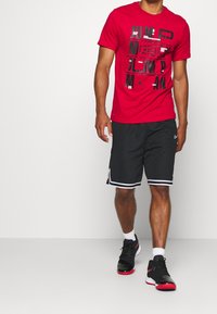 Rode graphic t-shirt met zwarte "Jumpman" tekst, gecombineerd met zwarte shorts met witte strepen en zwarte sneakers met rode accenten.