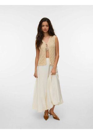 Femme aux longs cheveux bruns porte un top beige crochet sans manches, une longue jupe crème et des talons pointus marron, tenant le tissu de la jupe d'une main.