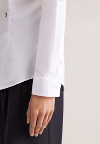 Lewa ręka ubrana w białą koszulę z długim rękawem, zapiętym mankietem na guziki i małą czarną haftowaną różą, spoczywająca obok czarnych spodni.