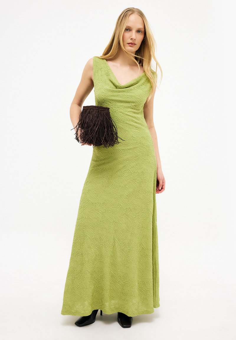 Naine, kes kannab tekstuurse mustriga helesinist varrukateta maksikleit, musti pahkluu-saapaid ning hoiab käes tumepruuni lukkudega väikest käekotti, taust on ühevärviline.