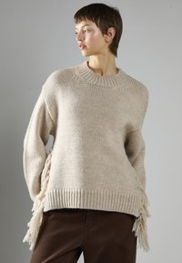Młoda osoba w beżowym swetrze dzianym z frędzlami po bokach i ciemno-brązowych spodniach, patrząca w lewo na neutralnym tle.