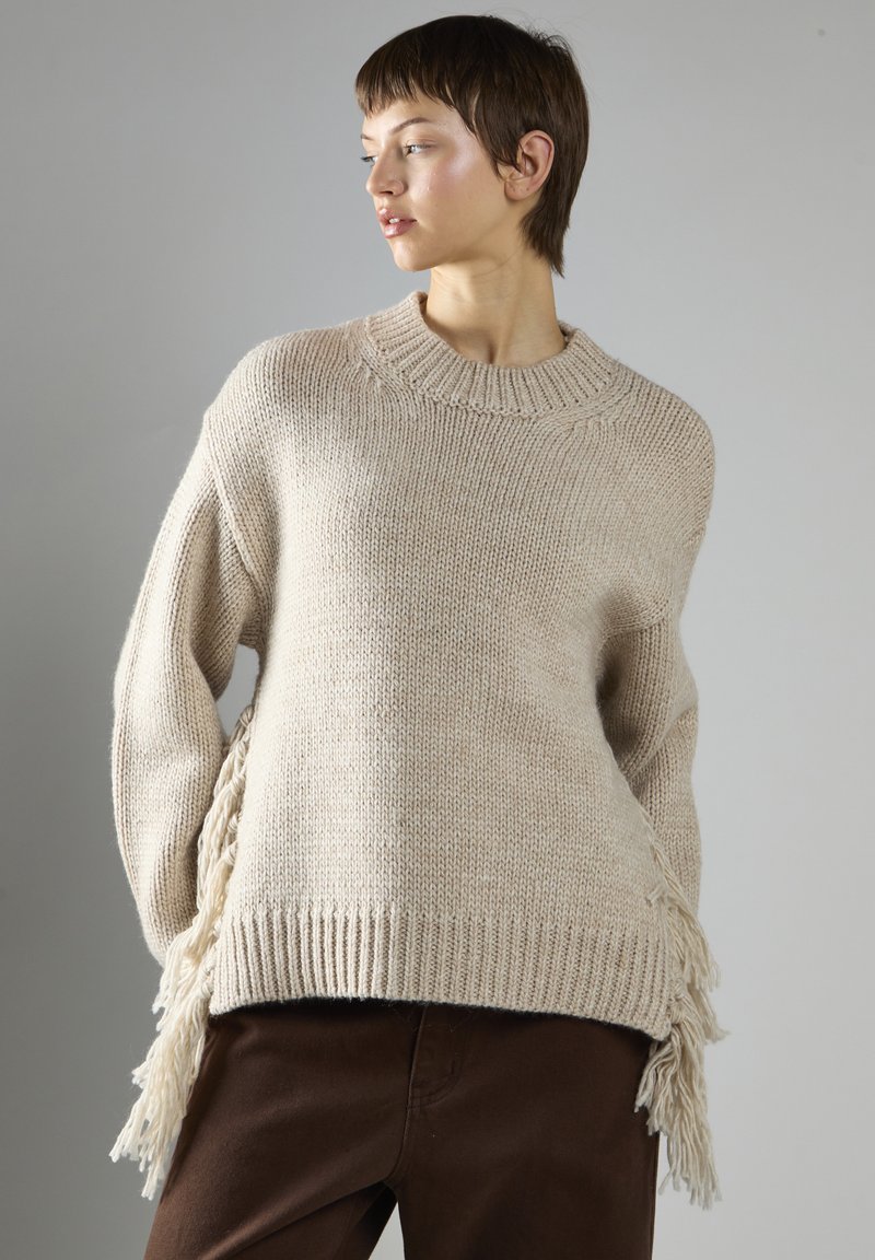 Młoda osoba w beżowym swetrze dzianym z frędzlami po bokach i ciemno-brązowych spodniach, patrząca w lewo na neutralnym tle.