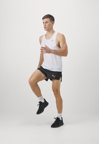 Biały bezrękawnik sportowy o subtelnej fakturze, połączony z czarnymi szortami. Ubranie ma małe logo i buty biegowe.