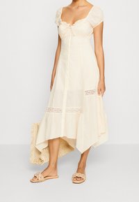 Woman wearing cream midi dress with lace inserts, short puff sleeves, button-down front, holding woven straw bag, and beige braided slip-on sandals.