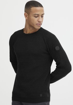 Black knit sweater with a textured pattern, ribbed neck, and cuffs. Features a circular branding patch on the left sleeve.