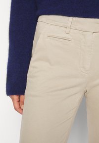 Beige Hose mit seitlichen Taschen, kombiniert mit einem dunkelblauen Langarmpullover, der einen Teil der Hand- und Taillenpartie zeigt.