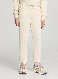 Pantaloni della tuta beige chiaro con elastico in vita e polsini alla caviglia, realizzati in tessuto morbido. Abbinati a sneakers beige con design a motivi.
