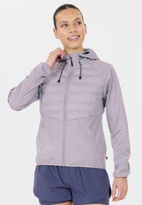 Kvinde justerer hætten på en lys lilla zip-up jakke, iført mørkeblå shorts, smilende og ser til siden på en hvid baggrund.
