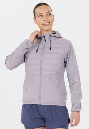 Vrouw die de capuchon van een lichtpaarse zip-up jack aanpast, gekleed in marineblauwe shorts, lachend en naar de zijkant kijkend op een witte achtergrond.