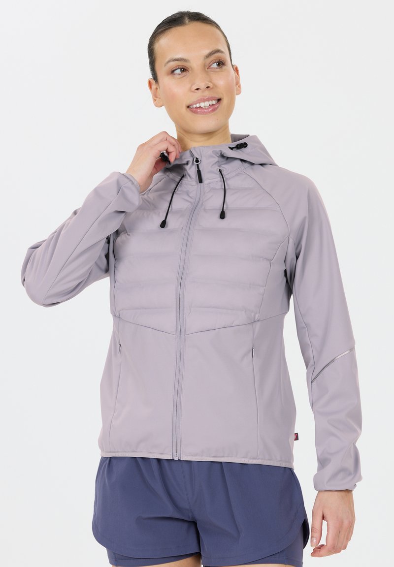 Kvinde justerer hætten på en lys lilla zip-up jakke, iført mørkeblå shorts, smilende og ser til siden på en hvid baggrund.
