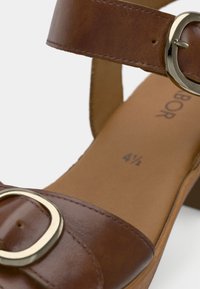 Καφέ δερμάτινο σανδάλι με φαρδιά λουρί, στρογγυλεμένη μύτη, μεγάλο αγκράφα σε χρυσή απόχρωση και ανοιχτό καφέ υφασμάτινο εσωτερικό με τυπωμένο μέγεθος παπουτσιού.