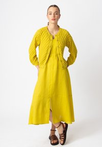 Kollane tikitud kardigan ümara kaelusega, nööpide sulgemise ja pikkade varrukatega; koos sobiva kleidiga, millel on ees lõhik ja looduslik kangas.