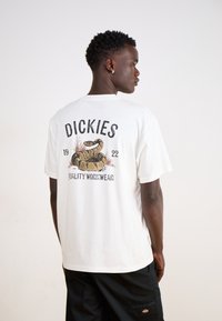 White short-sleeve t-shirt with a back graphic. Features a snake design, brand name "Dickies," and "Quality Workwear" text. Cotton material.