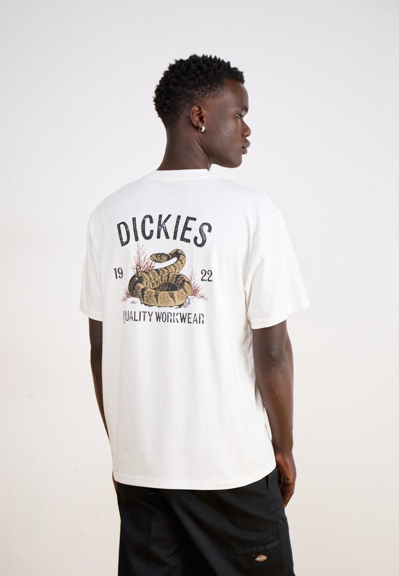 Camiseta blanca de manga corta con un gráfico en la parte posterior. Presenta un diseño de serpiente, el nombre de la marca "Dickies" y el texto "Ropa de trabajo de calidad". Material de algodón.