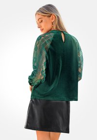 Groene fluwelen top met doorschijnende kantmouwen, keyhole-detail aan de achterkant en een losse pasvorm, gecombineerd met een zwarte leren rok.