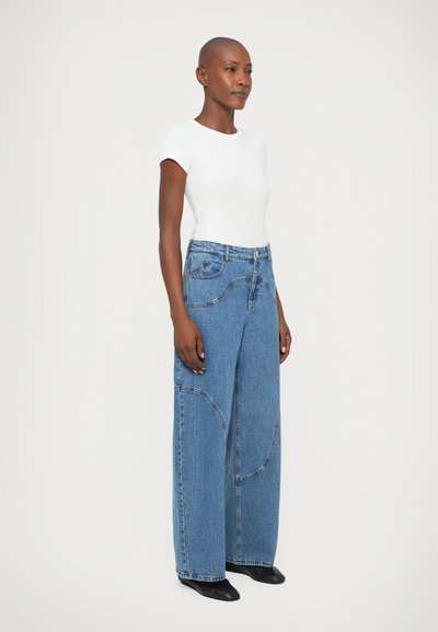 Bredbyxorna i ljusblå denim har synlig sömnad och hög midja, kombinerat med en åtsittande vit topp. Slät textur, avslappnad passform.