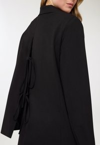 Blazer nero su misura con colletto alto, davanti aperto e schiena dettagliata con laccetti neri coordinati e una texture liscia.