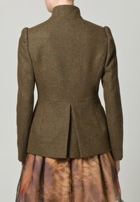 Mujer vistiendo una chaqueta de tweed marrón ajustada con hombros estructurados y una espalda acampanada, combinada con una falda transparente y con estampado.
