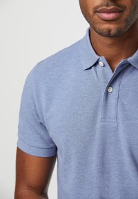 Světle modrá polo košile vyrobená z texturované tkaniny. Má límeček s dvěma bílými knoflíky a krátké rukávy.