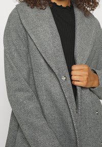 Donna che indossa un cappotto grigio a motivo rigato con ampi risvolti sopra un top a coste nero, tenendo chiuso il cappotto.