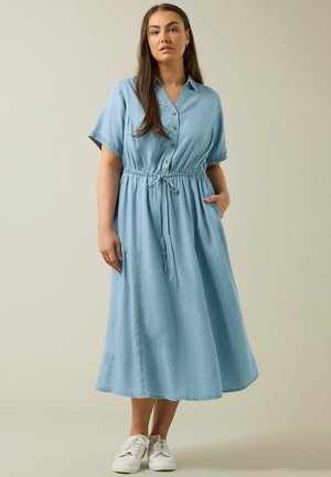 Vrouw staand, draagt een lichtblauwe midi-jurk met knopen en korte mouwen, en witte sneakers, één hand in de zak, neutrale achtergrond.