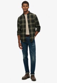 Groene geruite flanellen shirt over een lichtbeige t-shirt, gecombineerd met blauwe spijkerbroeken en bruine veterschoenen. Minimale hardware, casual pasvorm.