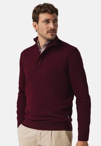 Bordeauxroter Pullover mit hohem Kragen und Knopfakzenten, aus weichem Stoff; kombiniert mit hellfarbenen Cordhosen.