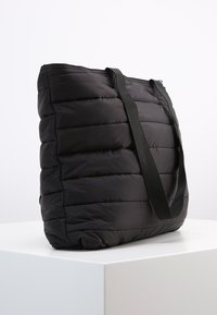 Sac fourre-tout noir matelassé, avec une texture matelassée, doté de deux longues poignées noires et d'un design spacieux. Extérieur en tissu lisse, détails minimaux.