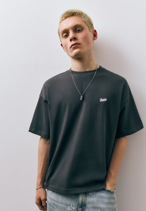 Ung person med kort blondt hår iført en mørk oversized t-shirt, lyse denimjeans med hvid broderi, sølvhalskæde og armbånd.